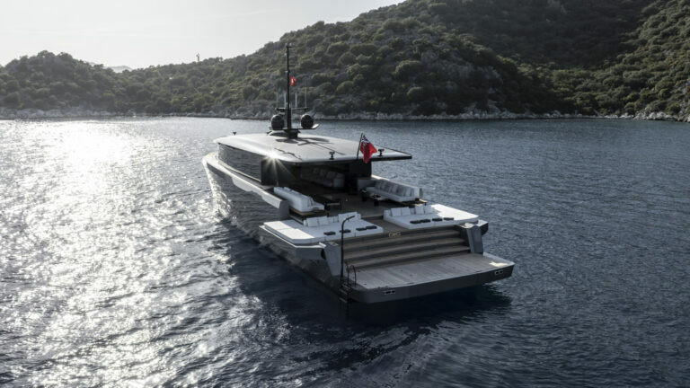 Elevated view of a large luxury yacht anchored in a calm bay, with a rear platform extended over the water. The yacht's deck is outfitted with sunbeds and a lounge area. The water glistens under the sunlight, and the vessel is surrounded by a lush, hilly landscape. The yacht's modern design features clean lines and a dark hull, with a distinctive flat-top structure that carries two flags proudly fluttering in the wind