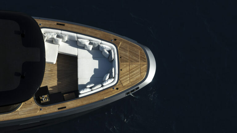 Aerial view of the bow section of a luxury yacht showcasing a curved wooden deck with built-in seating covered in white upholstery. The seating is arranged in a U-shape, creating an intimate lounge area. The yacht is surrounded by dark blue ocean waters, and sunlight creates shimmering reflections on the surface, highlighting the contrast between the natural wood tones and the vessel's sleek, dark exterior.